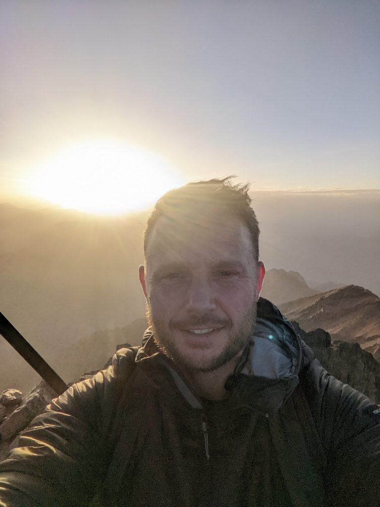 Tom standing at the peak of Mount Toubkal with the sun rising behind, after climbing mount toubkal